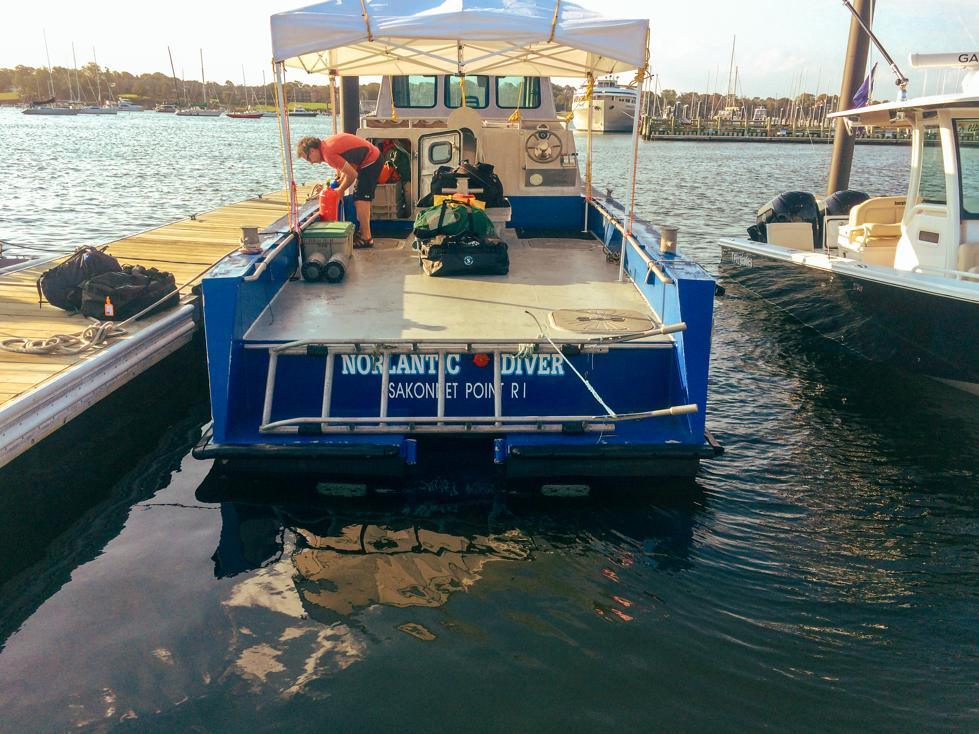 A man is standing on the deck of a blue dive boat unloading diving gear onto a wharf in Newport Harbor 